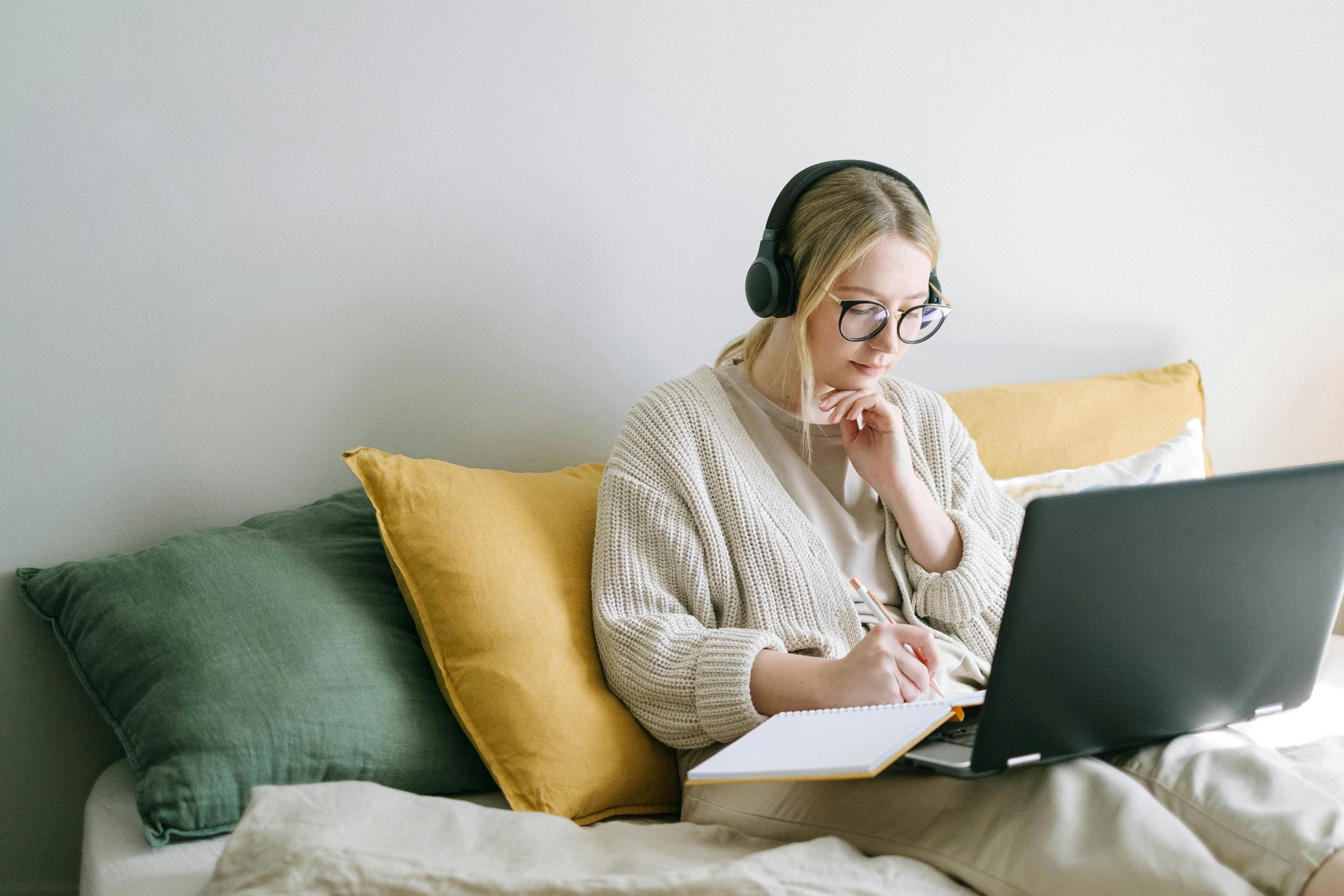 A focused woman in glasses and headphones works on a laptop from a cozy bed.