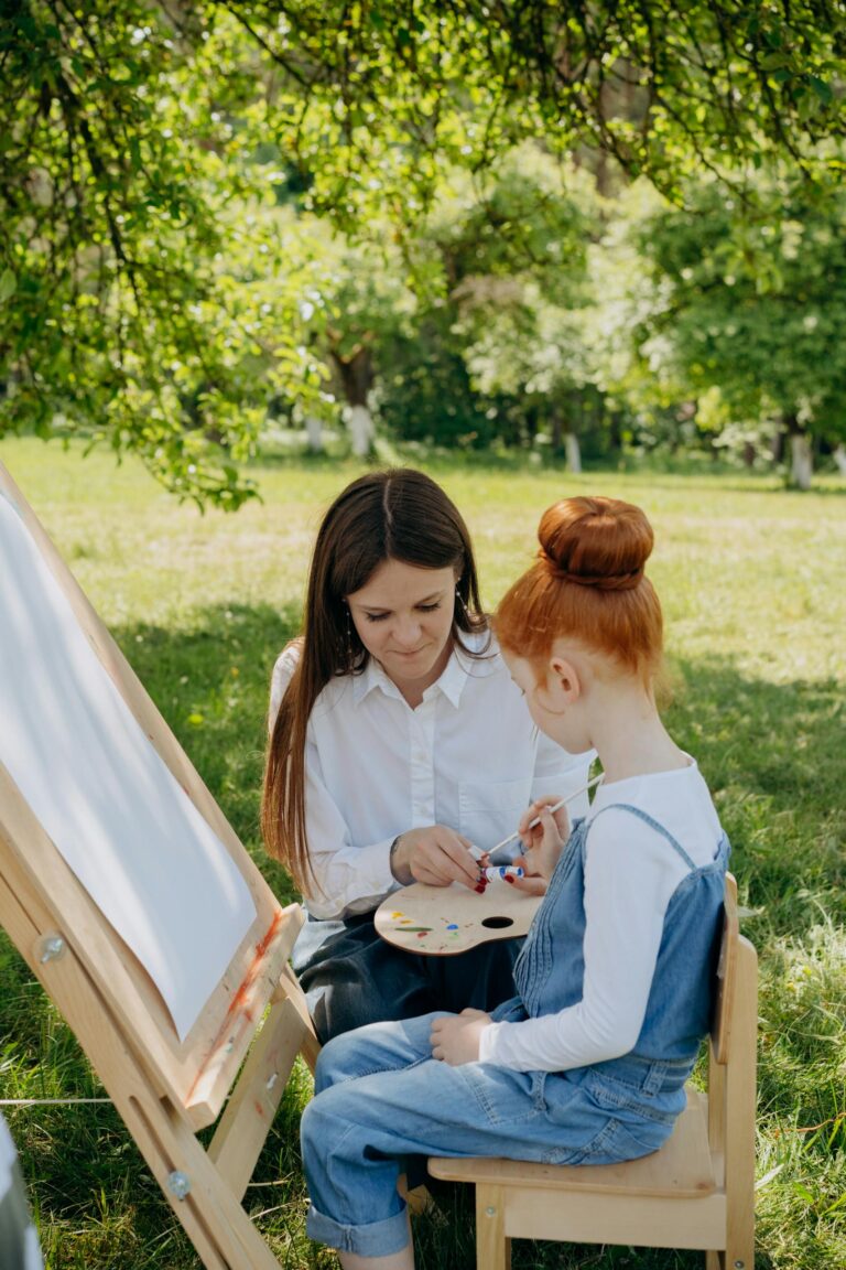 A young girl learning painting with her teacher in a sunny garden setting.